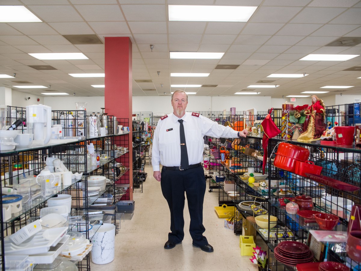 A man dressed in a shirt, pants, and a tie stands in the aisle of a thrift store. On the shelves around him, a variety of items, including sets of dishes, angel decorations, pots, pans, a faux-marble trash can, silverware drawer organizers, and more.