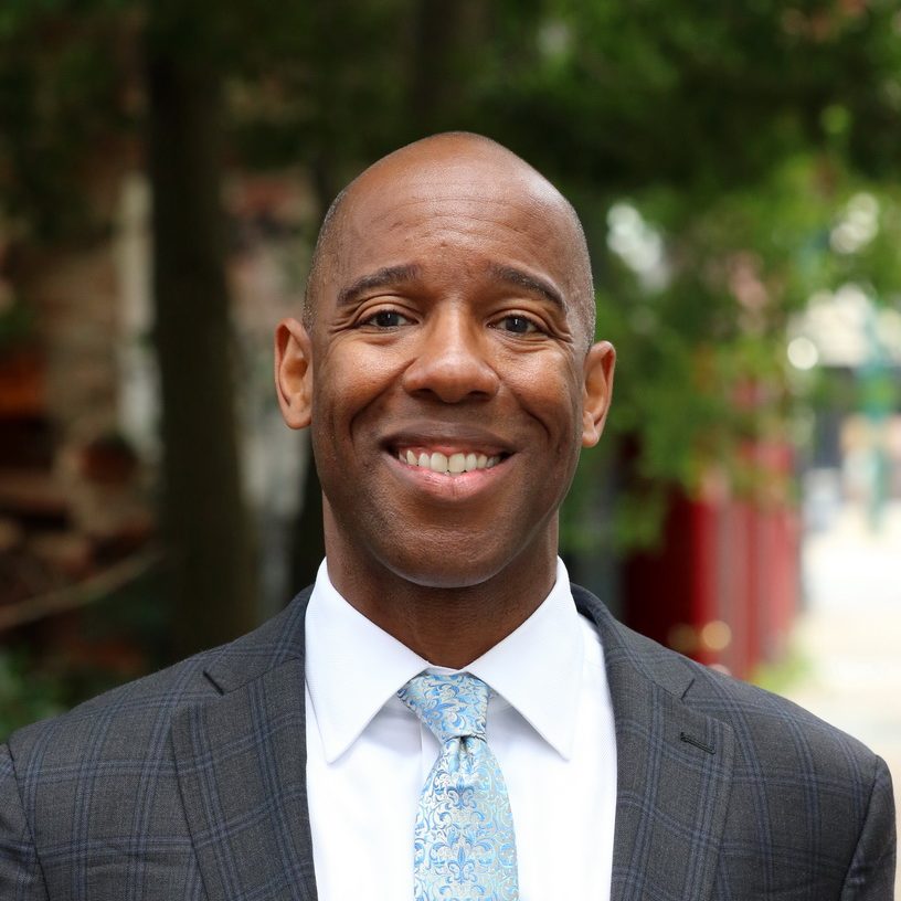 Man in a grey suit jacket and light blue tie smiling at camera, trees in background