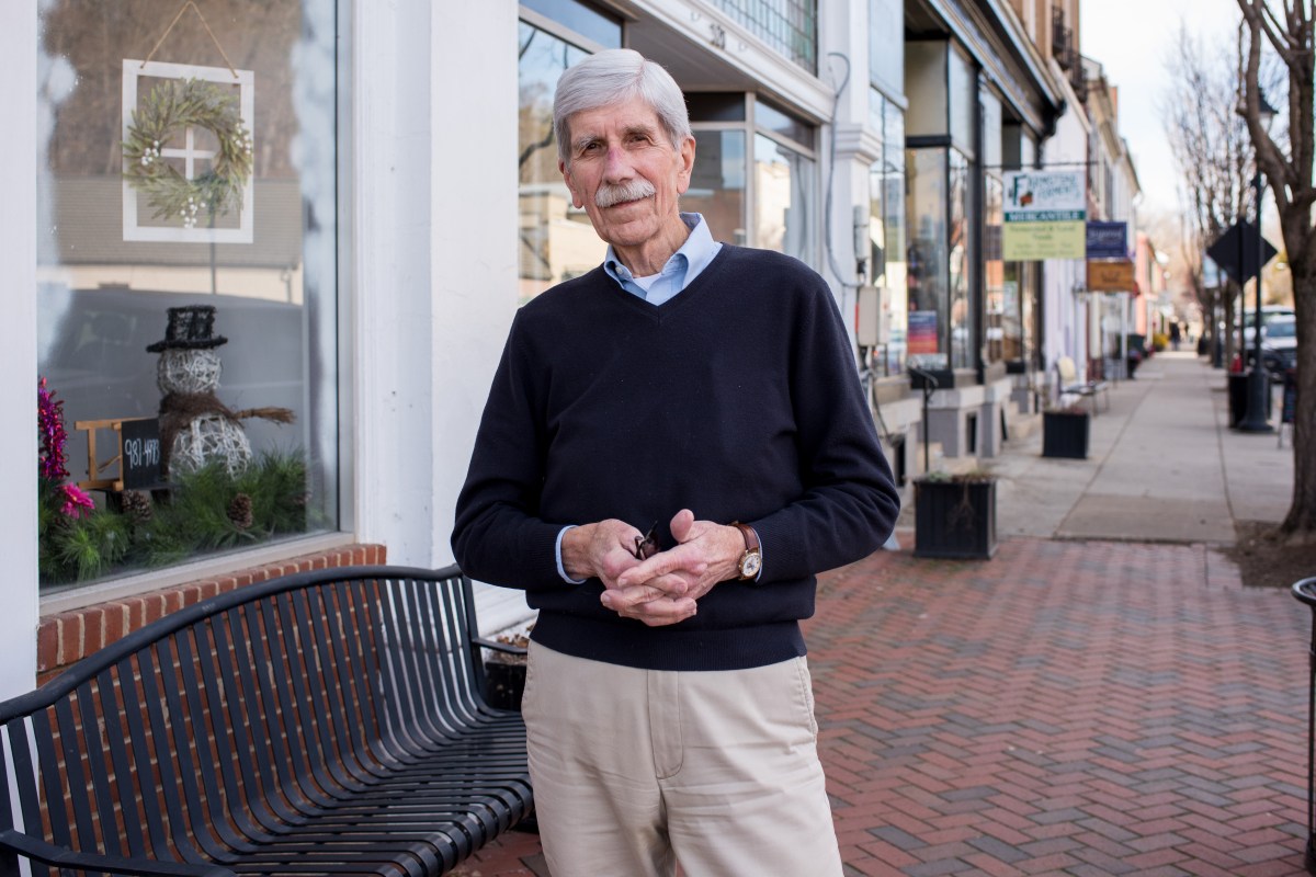 A man stands on a small town main street, shop windows and shop signs visible in the distance behind him. He is looking at the camera, and his hands are intertwined in front of him. He is holding a pair of glasses in his hands.