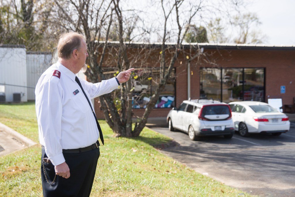 A man points toward a single store brick building with cars parked in front of it.