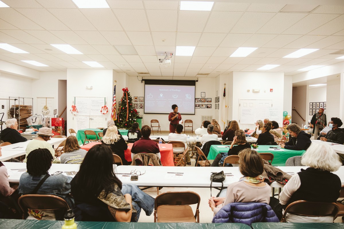 A brightly-lit room, with folding tables covered with Christmas-colored table cloths. A couple dozen people sit in folding chairs, watching a presentation being given at the front of the room. At the front of the room, a woman stands in front of a projector screen holding a microphone and talking to the audience. There are holiday decorations in the room, like a decorated tree, some wreaths, and balloons.