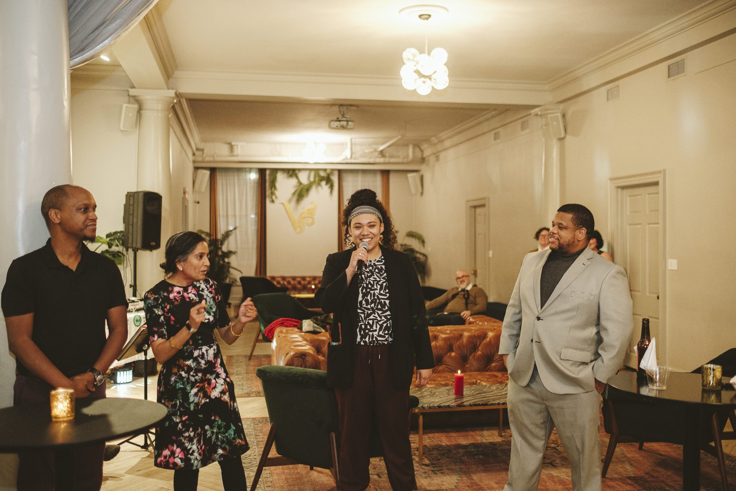 Four people standing in a stylish room, engaged in conversation. A man in a suit smiles, while a woman holds a microphone. Two others stand nearby, one clapping. Cozy seating and decor are visible.