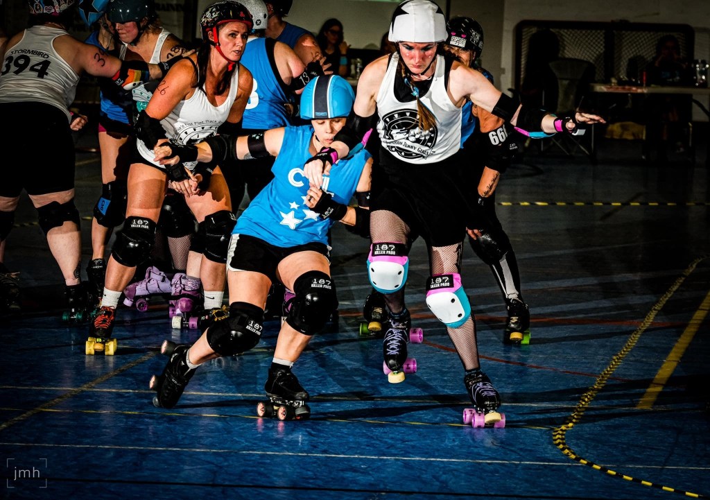 Two people in helmets, elbow pads, knee pads and roller skates bump into each other on a blue track. Behind them are other people wearing helmets, pads and riding roller skates.
