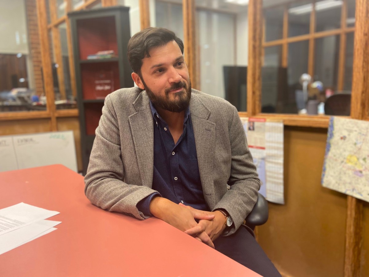 A person in a sport coat sits at a table with an office behind him.