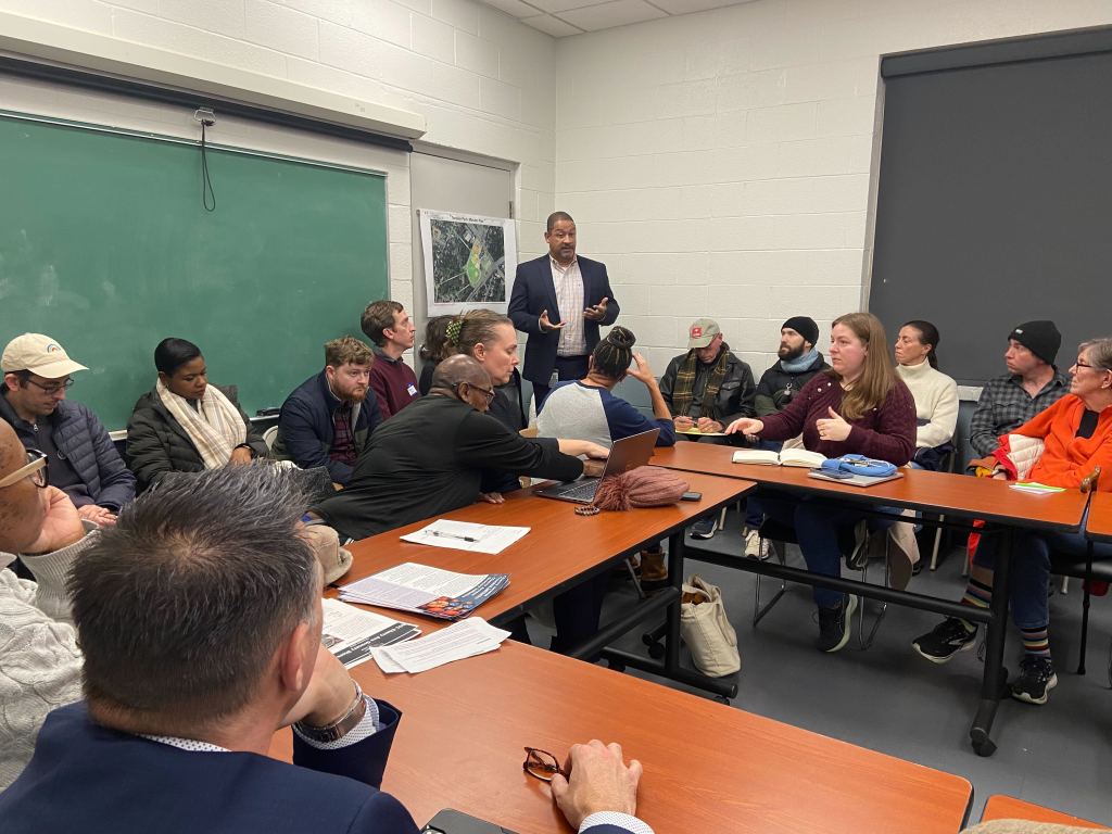 A few rectangular tables are set up end to end to form a squared-off circle. People are sitting at the tables, and against the walls, looking at a man who is standing up and speaking.