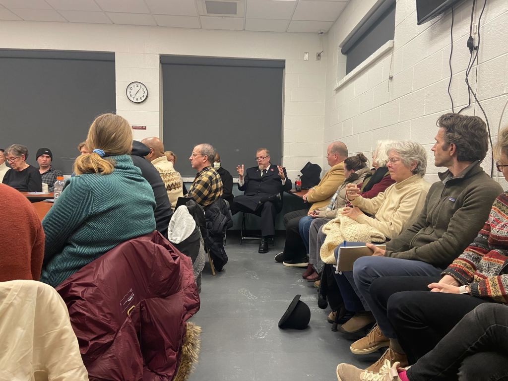 A group of people pack into a room with a bare concrete floor and walls.