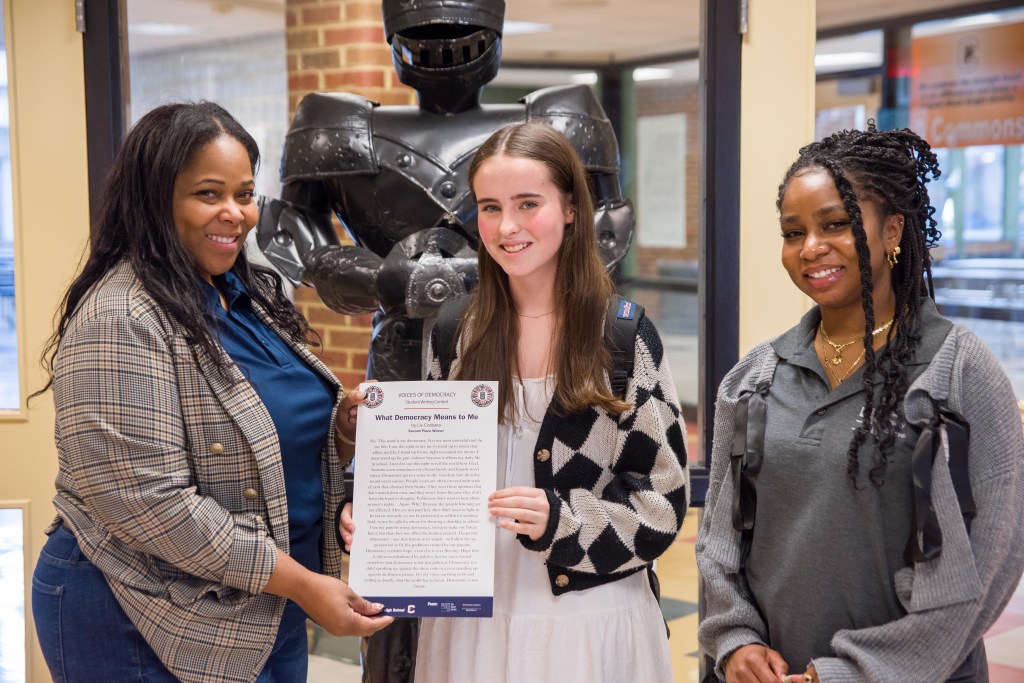Three people stand indoors next to each other, smiling and holding a document with an essay written on it. Behind them is a statue of a suit of armor and large windows.
