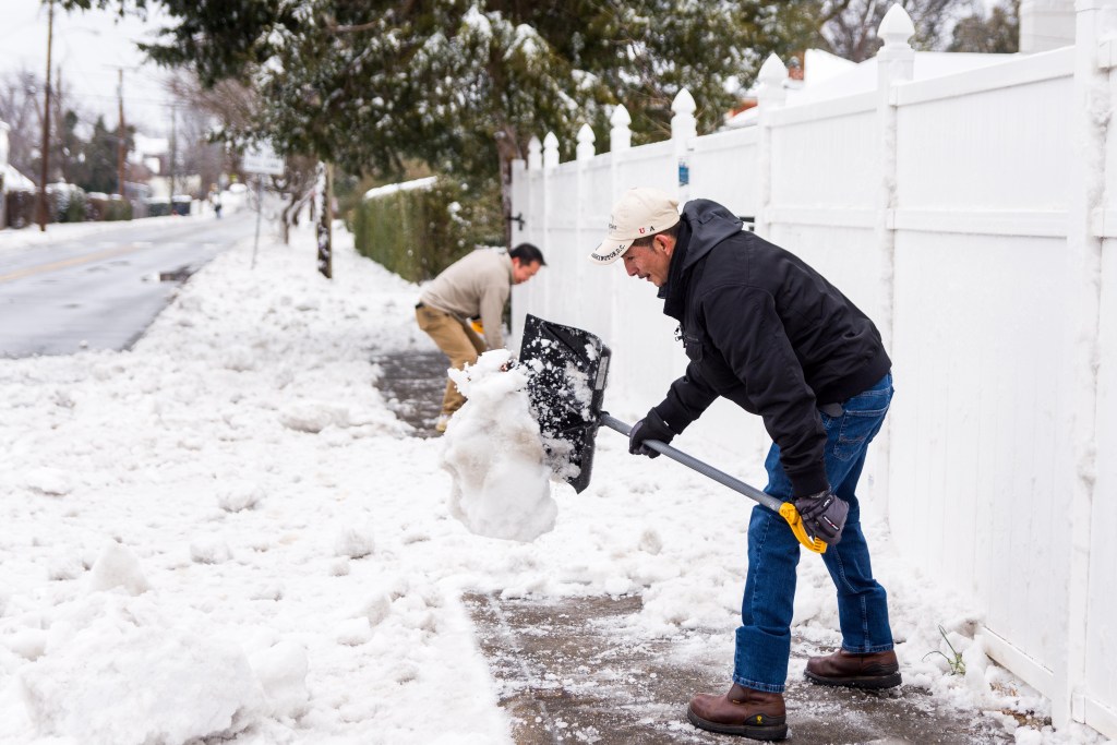 Two people shovel snow off a sidewalk, with a tall, white fence behind them.
