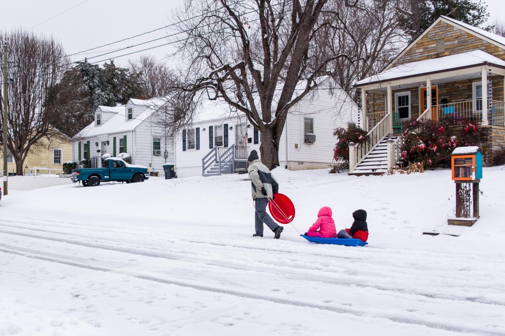 A residential street with three homes in view, an adult walking down a snow-covered street, pulling two children in snow gear on a blue sled.