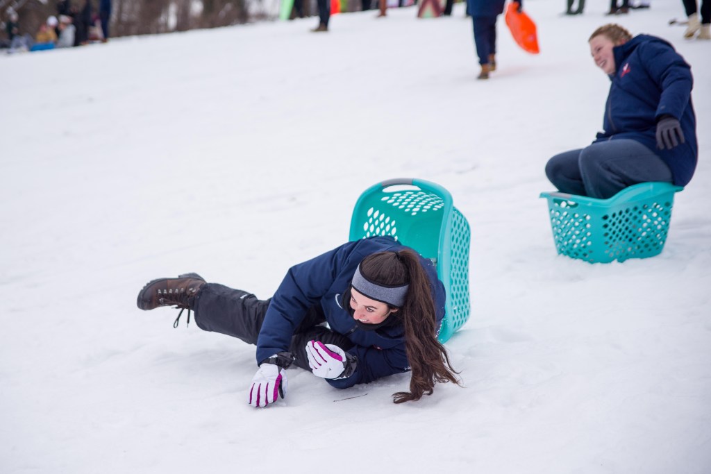 A woman in the snow falls out of a teal laundry basket, with a person in the background smiling.
