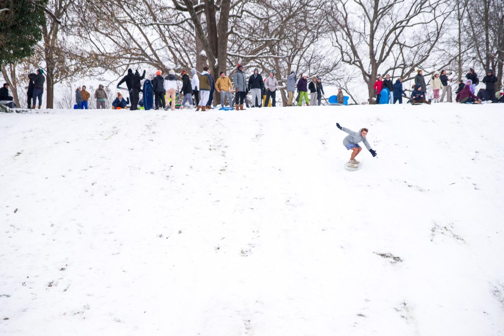 A line of people at the top of a hill watch a person on a board, in shorts, shooting down a snowy slope.