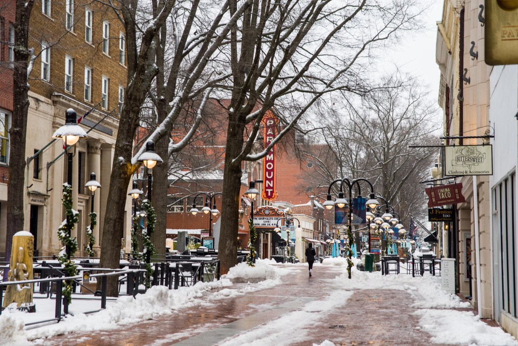 A street scene with a brick lane and edges covered in snow, a runner seen in the distance, trees and storefronts on either side.