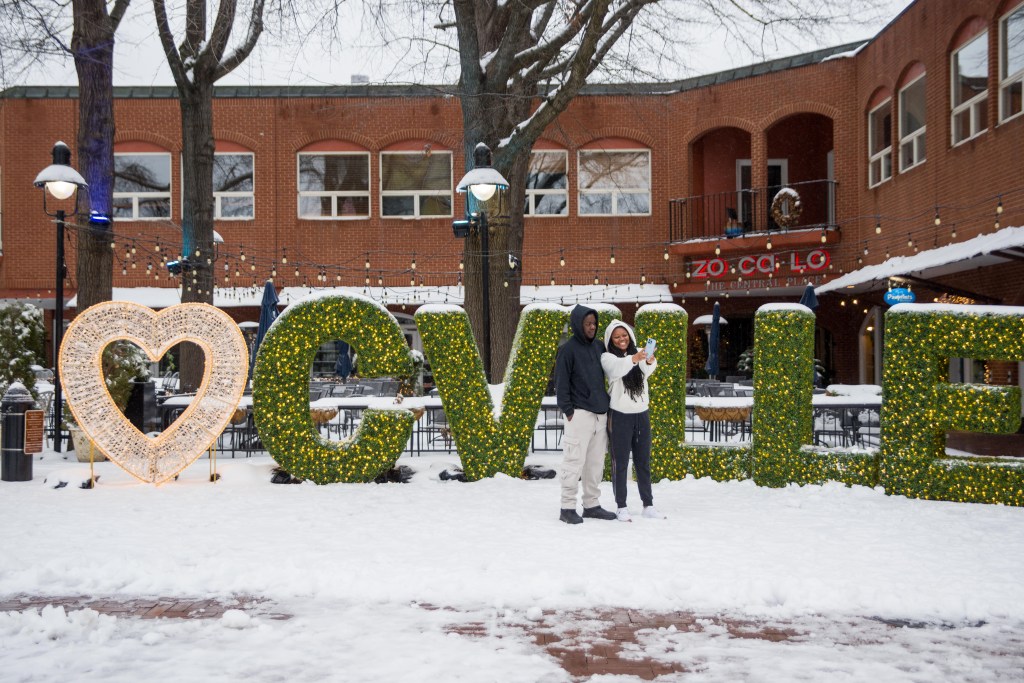 Two people take a selfie in front of a large installation with lights that has a heart and "CVILLE" in green, a brick building behind them.