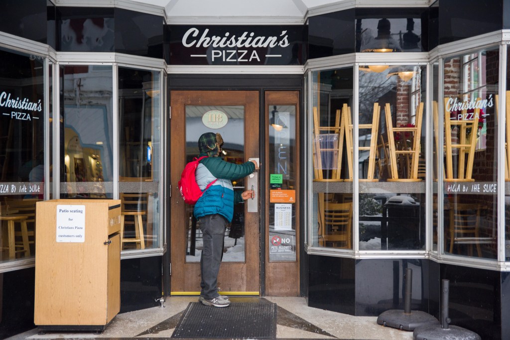 A person locking a door of a storefront with a sign that reads "Christian's Pizza" and bay windows with stools stacked on tables.