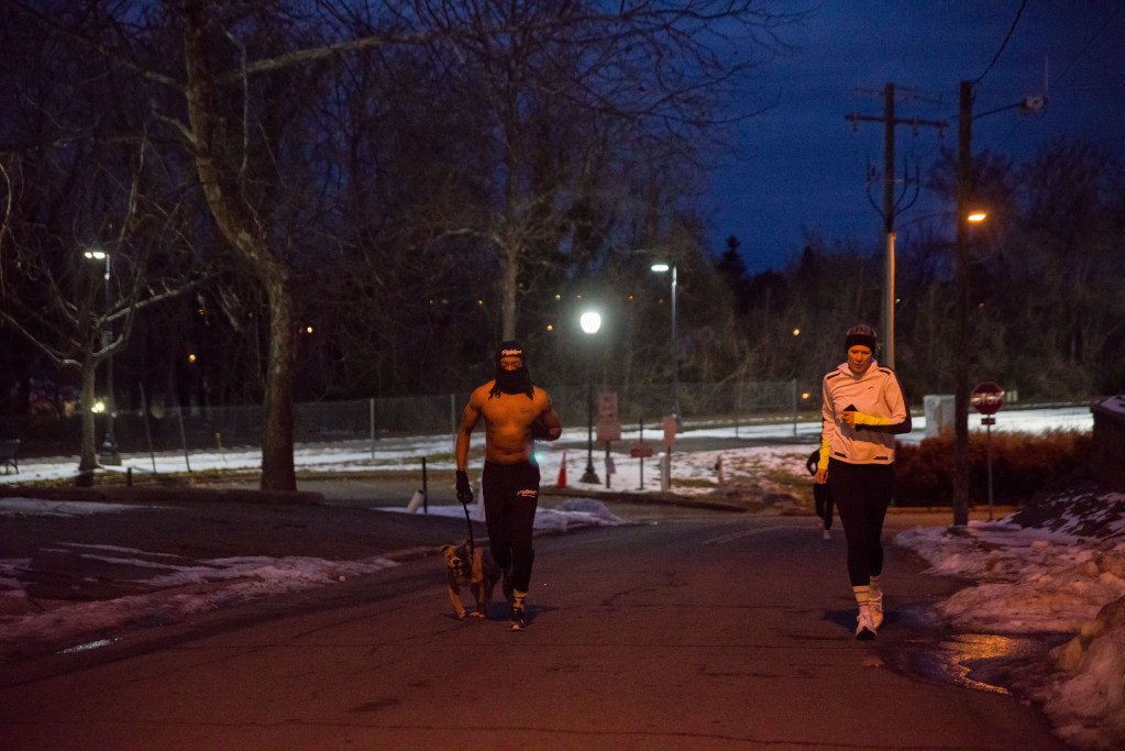 A man with a dog and a woman are jogging up a hill during the early morning. Snow is on the side of the running path. Another man jogging behind them can be seen at the bottom of the hill.