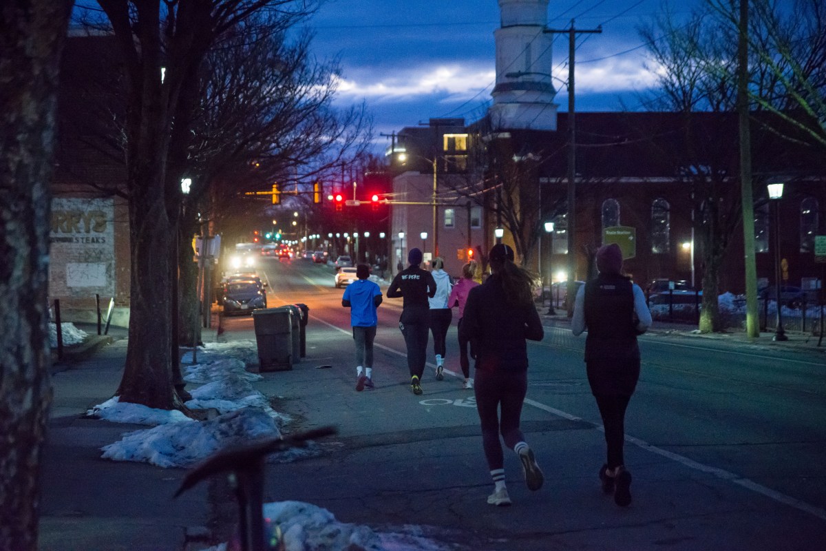 Six people are running down a street in the early morning amid a few cars on the road. Snow can be seen on the side of the street.