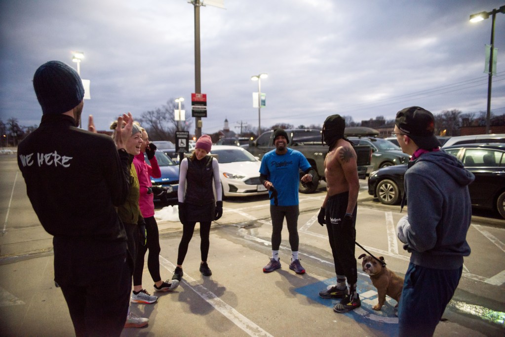 Seven people stand in a loose circle in a parking lot with cars behind them. They are wearing running clothes, running shoes, knit hats and gloves. One man in the circle is holding a dog on a leash.