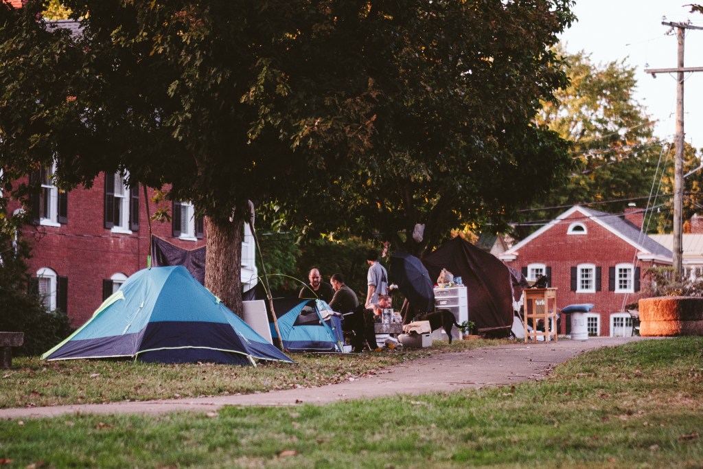 Three people and a dog are pictured at a distance in a city park surrounded by tents.