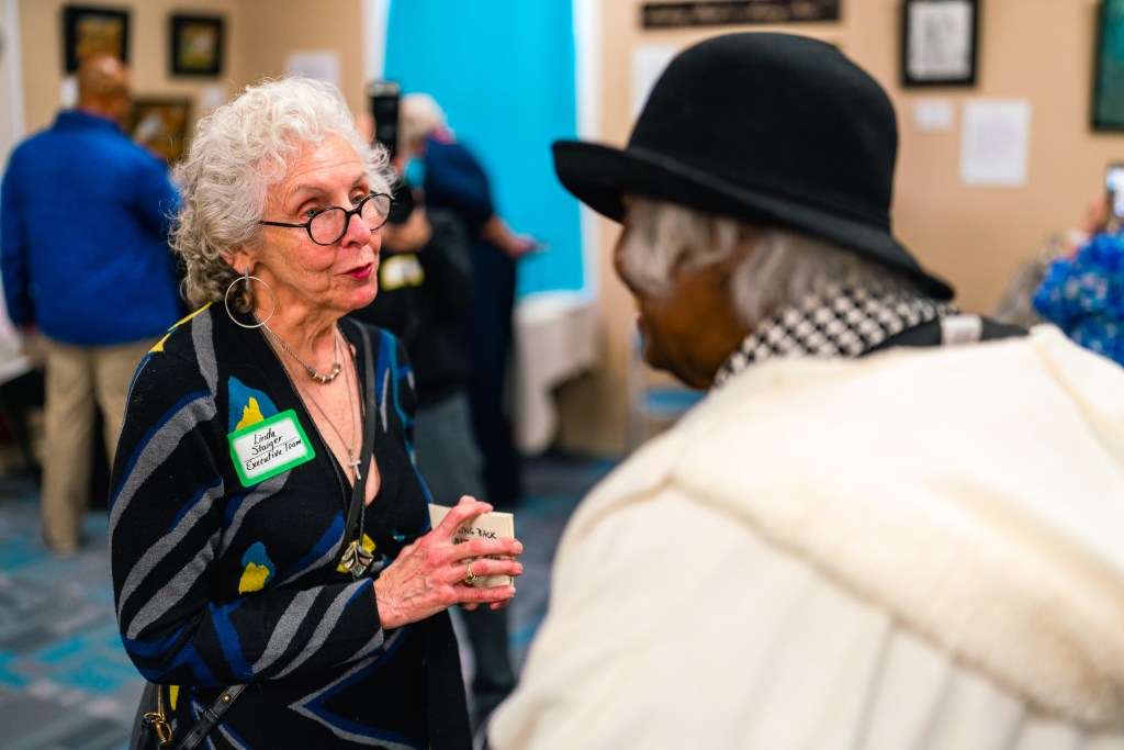 A woman talks to another woman while holding a card. Other people and pictures hanging on the wall are in the background.