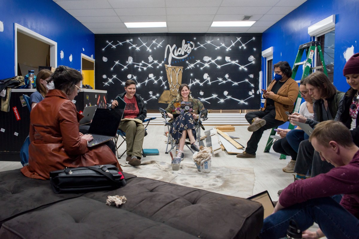 A group of nine people sit in a circle in a room on various chairs and couches facing each other, with brightly painted walls behind them.