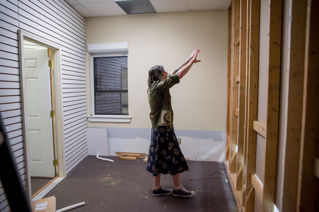 A woman stands in a room with bare walls, exposed framing and some construction materials scattered on the floor. She raises both her arms gesturing to an area against the right-hand wall.