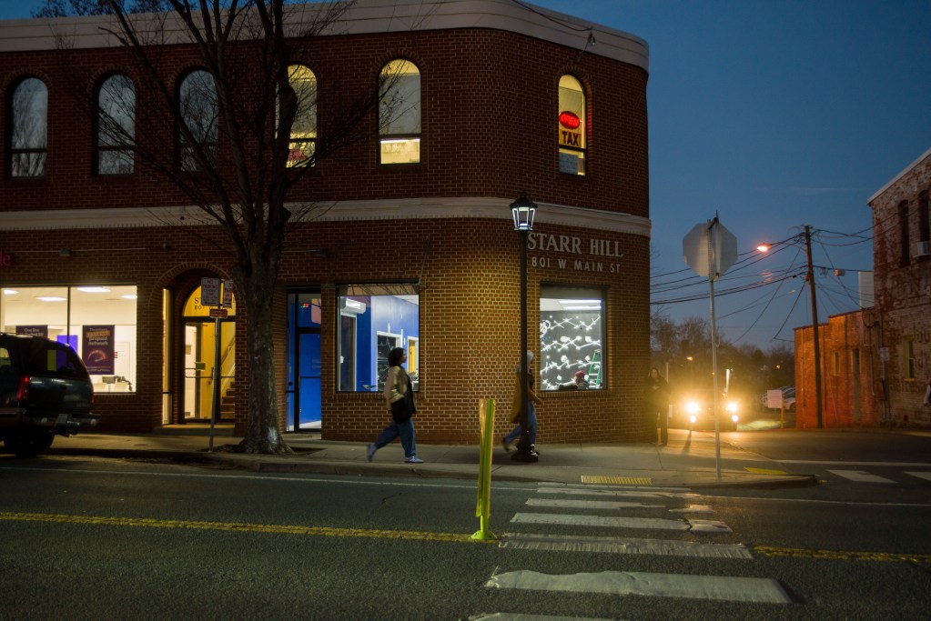A brick building is shown at night, with the words 'Star Hill, 801 W Main St.' above the corner window. Inside, people sitting in a colorful well-lit space as pedestrians walk by.