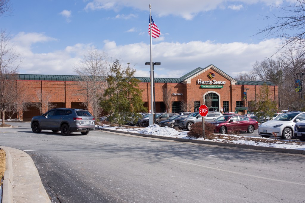 A brick building sits behind a salted parking lot filled with cars and small piles of plowed snow. A sign that reads, "Harris Teeter" hangs over the entrance.
