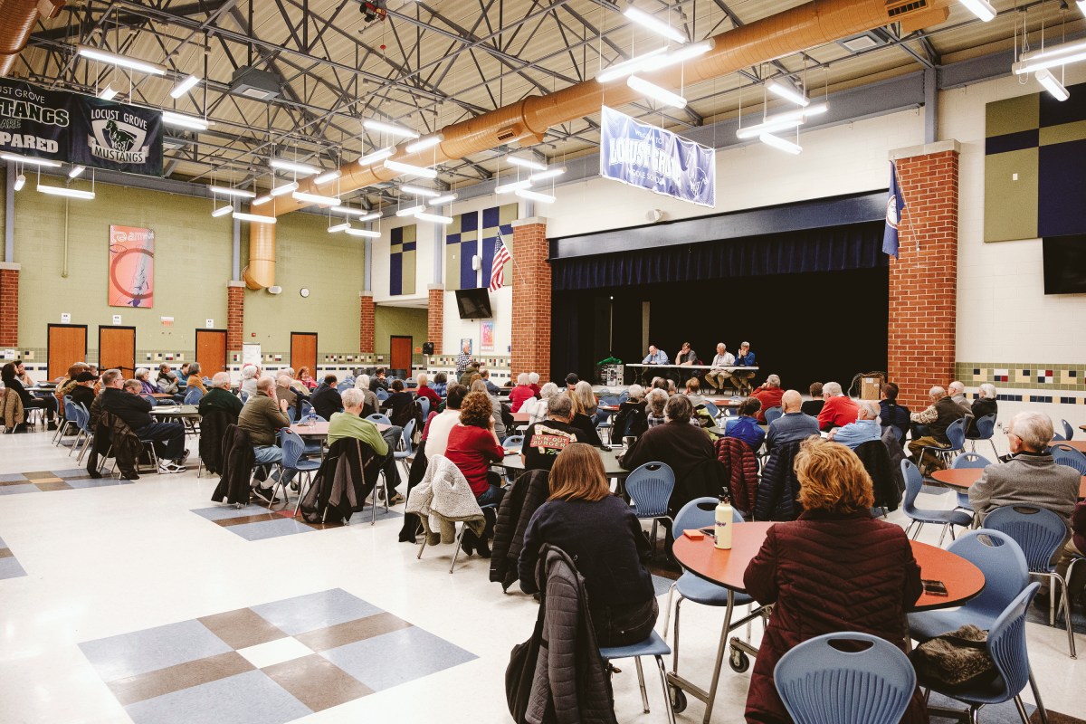 A large open space indoors where several people are seated around tables facing a stage. Four people are seated on the stage at a long table, facing the crowd.