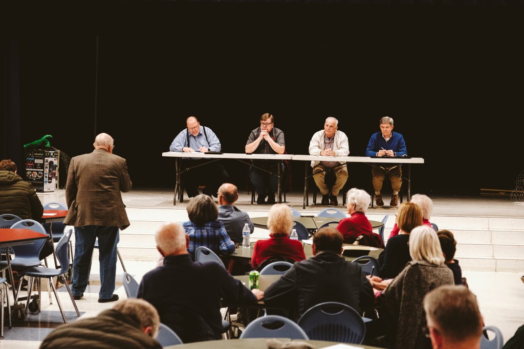 A man in a brown suit coat addresses a table of four people seated on a stage. Other people are seated around tables nearby.