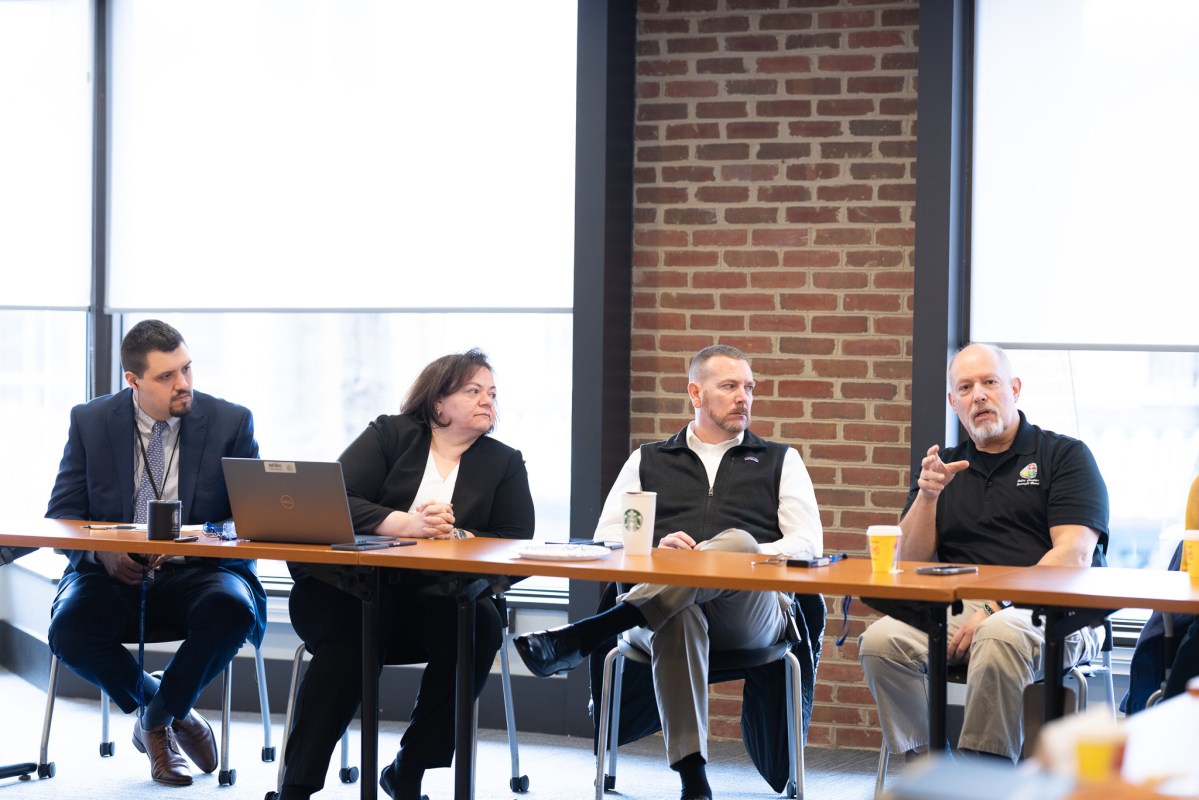 Four people dressed professionally sit at a long table. While one person speaks, gesturing with his hand, the other three watch him listening.