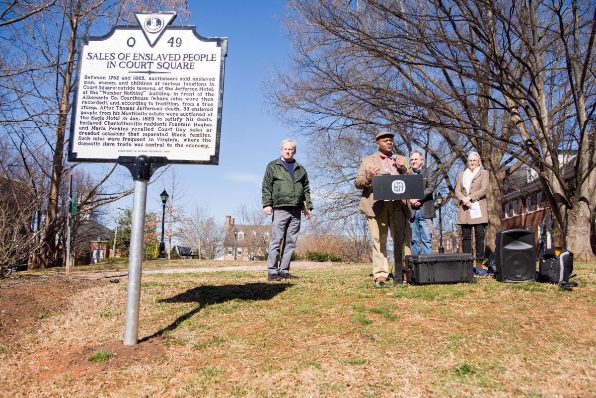 A historical marker reading 'Sales of Enslaved People at Court Square' is in the foreground. A man stands speaking at a podium with a microphone, with two more men and a woman standing on either side of him.