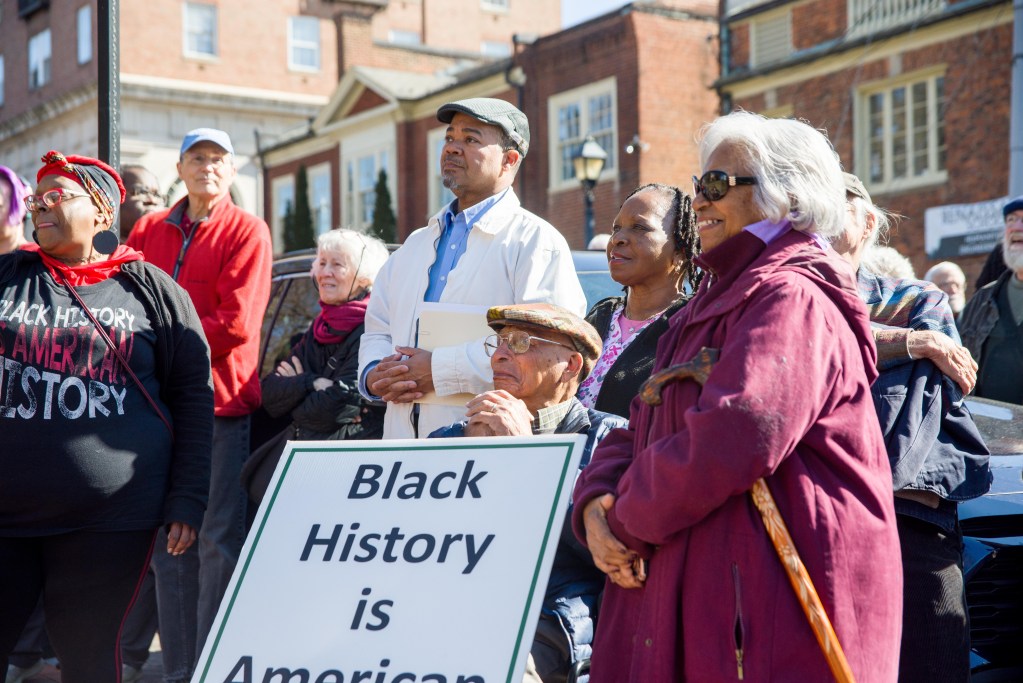 A crowd gathered outside, a man is seated at center with a large sign in front of him reading 'Black History is American History.' Other community members stand around him looking on; brick buildings can be seen in the background.