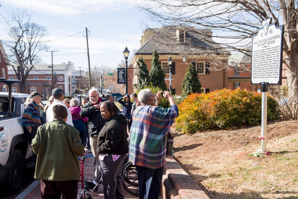 A crowd of people gather outside on the sidewalk during the day. On the right is a historical marker on a signpost with flowers laid at its base. People take photos, look at the marker and talk to each other.
