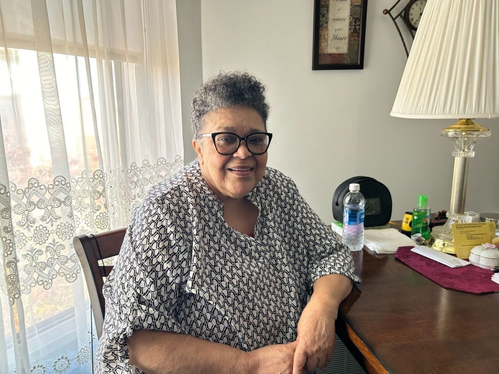An older woman with short, curly hair and glasses sits at a table, smiling.