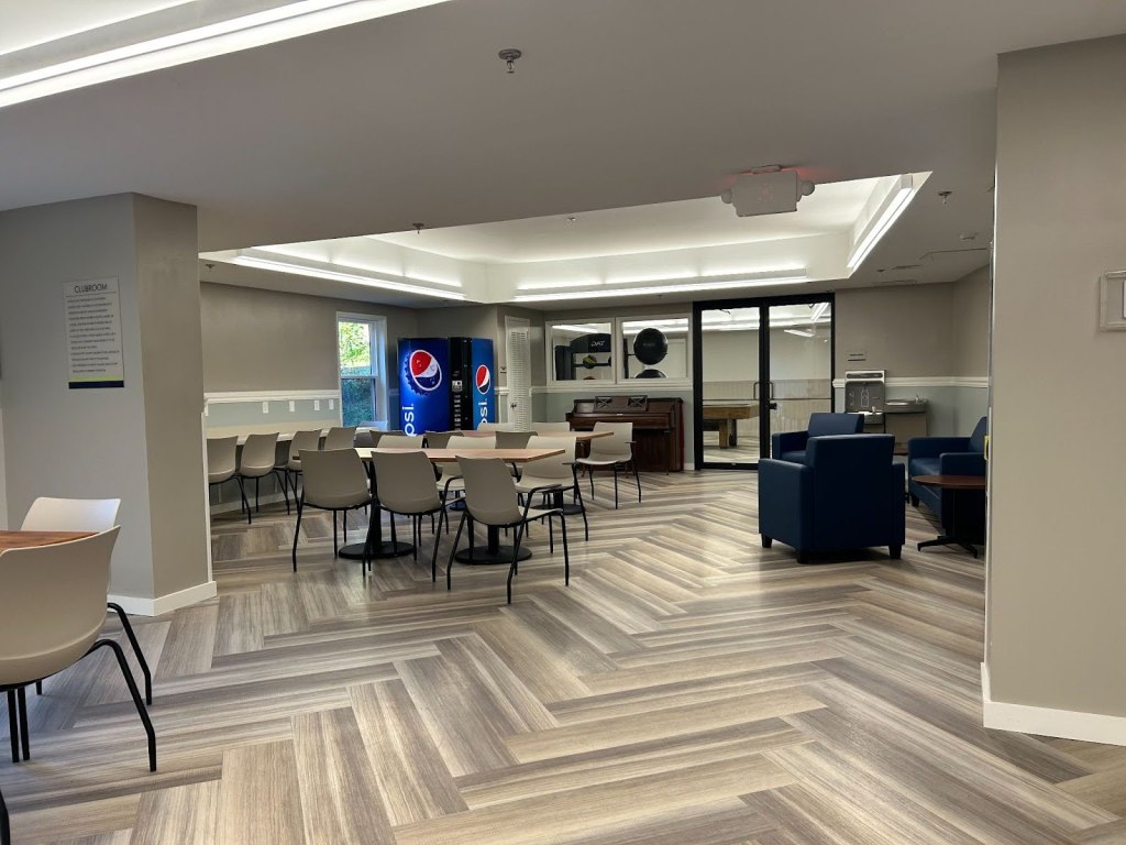 A photograph of a room. In the center, two tables surrounded by chairs. To the right, three armchairs and a coffee table. Along the back wall are a water fountain and a water bottle filling station, a piano, and a Pepsi vending machine. Visible through glass windows and doors at the back wall are a pool table and some exercise equipment.