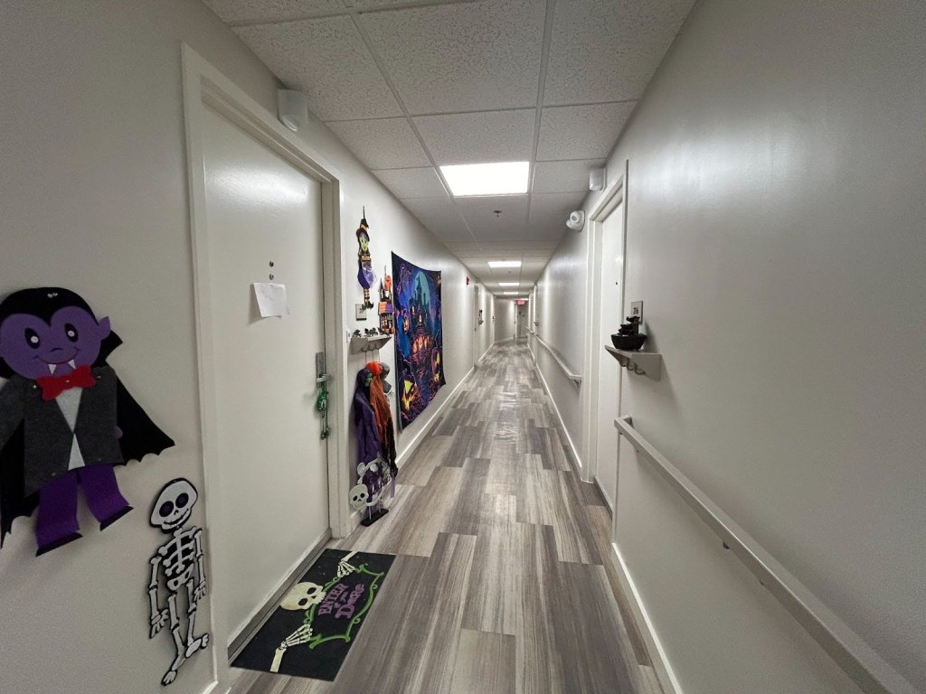 A photograph of a hallway, with new floors, new paint, new railings, new lighting, and new emergency lighting. The door to the left is flanked by Halloween decorations of vampires, skeletons, witches, and haunted houses.