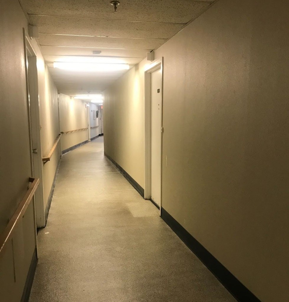 A photograph of a hallway. The floors, walls, and fixtures are worn. There is a light spot on the floor where a welcome mat used to be.