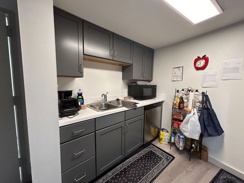 A photo of a small galley kitchen, with a double sink, cabinets, drawers, a dishwasher, and a microwave. There are narrow rugs on the floor, and a small metal tower with a can of coffee, chocolate sauce, mustard, paper towels, some reusable bags, and other items. There is an apple-shaped clock on the wall.