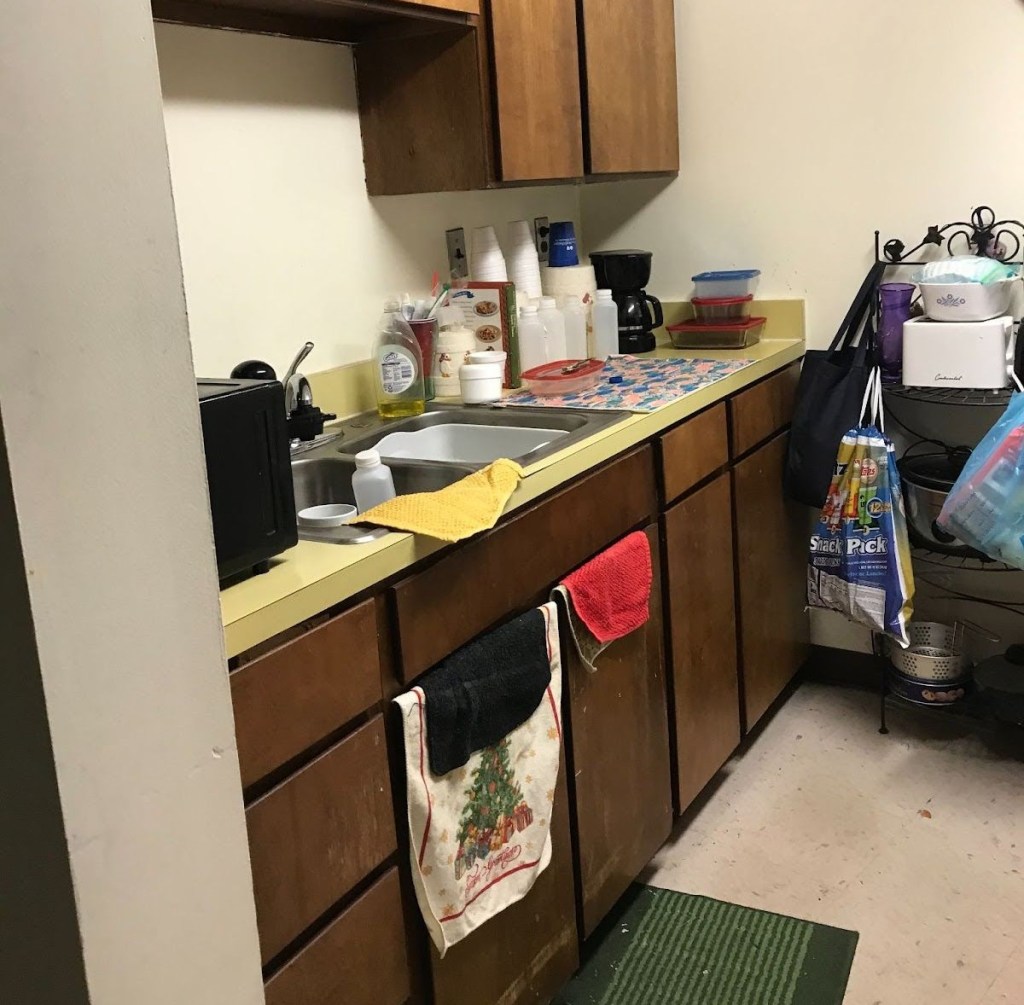 A view of a galley kitchen, with worn wooden cabinets and an old formica countertop. There are styrofoam cups, a few plastic food containers, plastic bottles, and a coffee pot on the countertop.