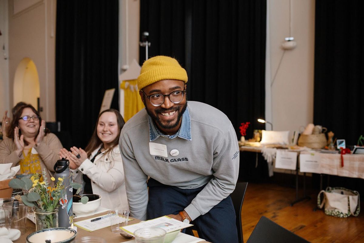 A man wearing glasses and a knit cap smiles as he stands up from his chair. In front of him is a dining table set with plates, glasses and floral arrangements. Other people are seated at the table applauding.