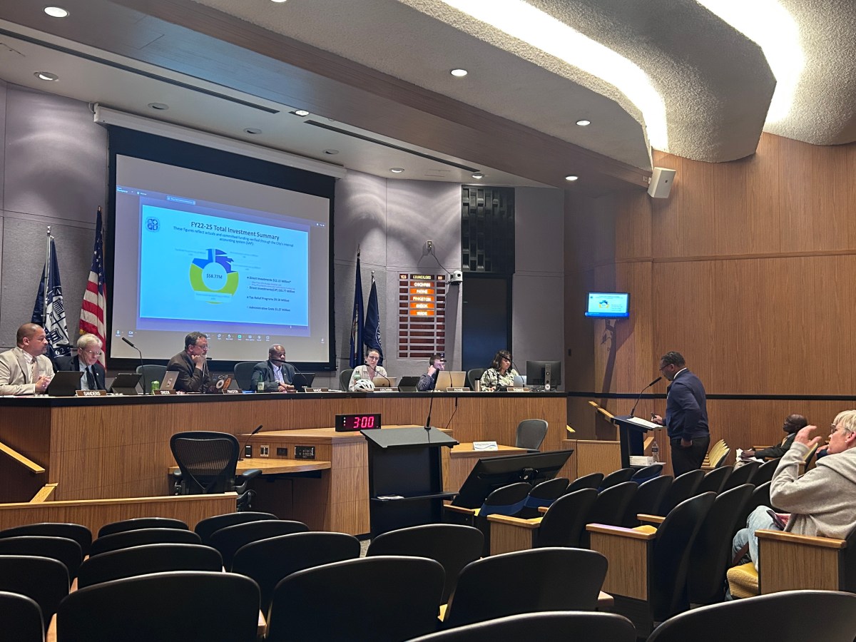 A photo of Charlottesville City Council chambers, with rows of chairs facing a dais. All five City Councilors, the City Manager, and the Clerk of Council sit at the dais, looking at a man in a suit who is speaking at a lectern. Behind the dais is a projector screen, with a presentation slide projected onto it.