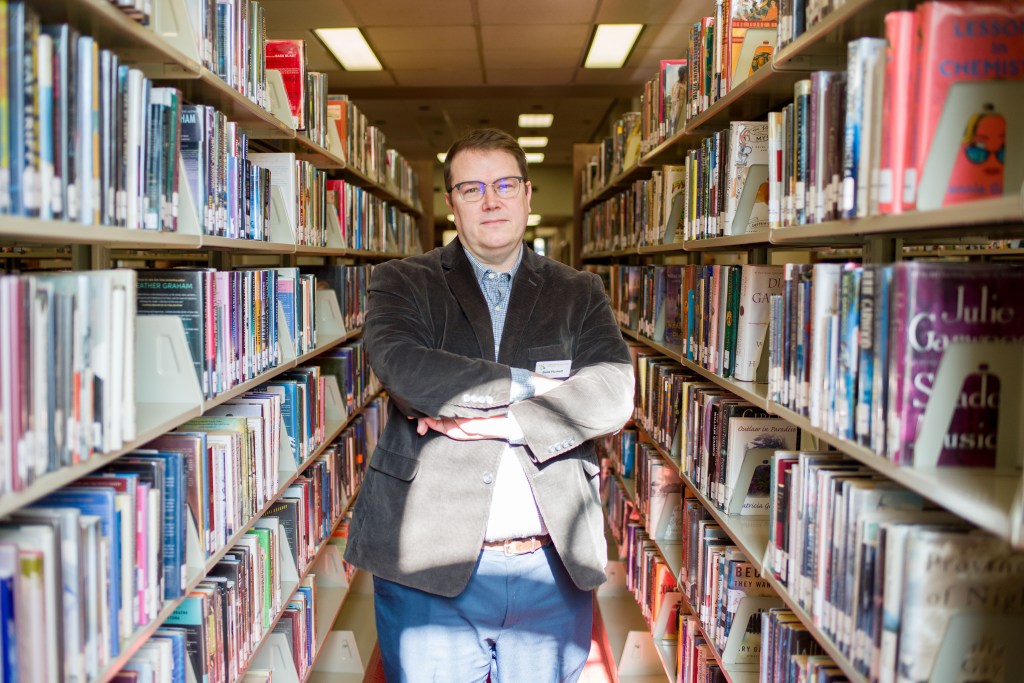 A man stands in library stacks, with long shelves of books on either side of him.