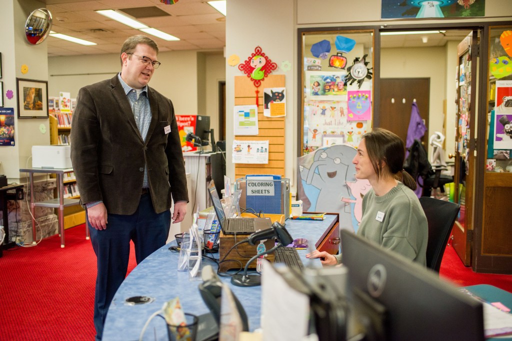 A woman sits behind a desk talking to a man who is standing. Colorful paper cutouts of children's book characters and original art by local kids decorate the walls behind them.