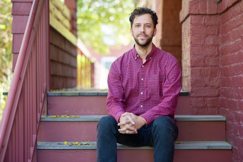 A man sits on a staircase outside of a house. He has a soft smile on his face, is resting his arms on his knees and clasping his hands.