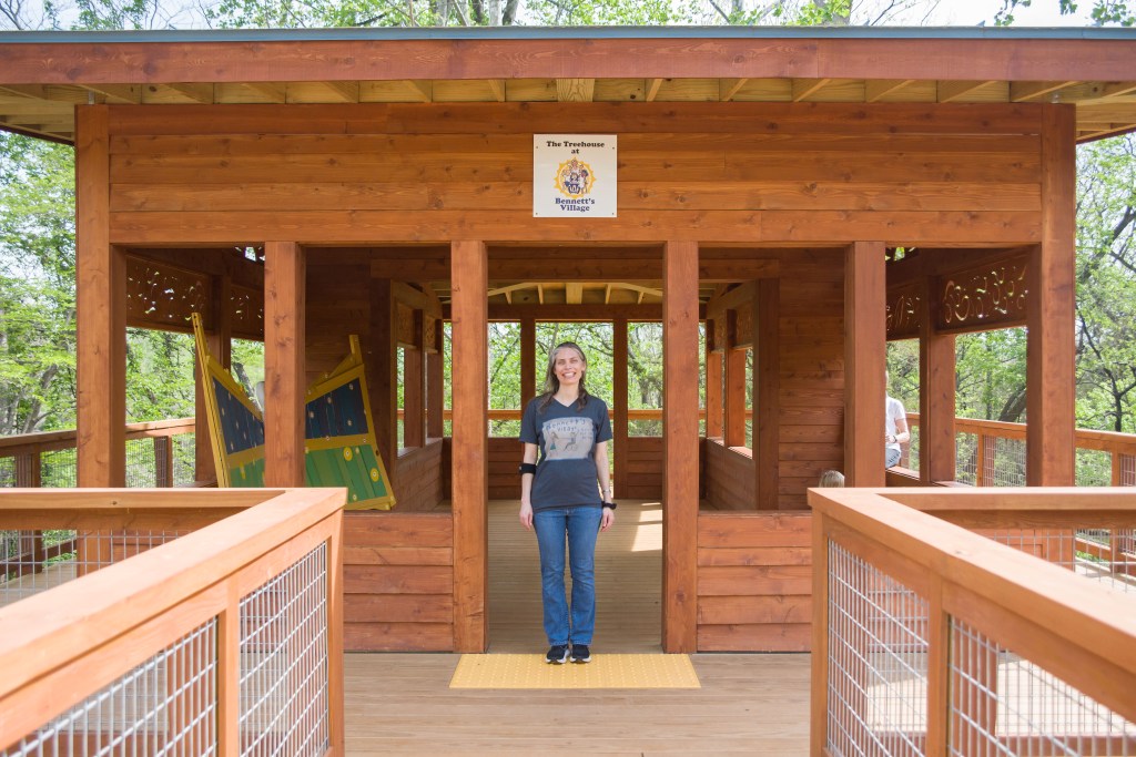 A woman stands in front of a large treehouse built of wood, with large trees visible in the background through the windows. A sign above the door reads 'The Treehouse at Bennett's Village.'