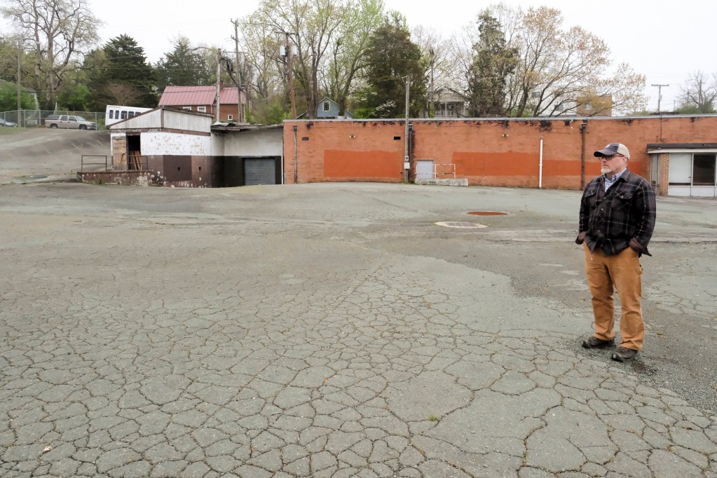 A photo of a long brick building with a sagging roof situated in a parking lot. The parking lot pavement is cracked. A man stands on the pavement with the building in the distance behind him. There are houses and trees in the background.