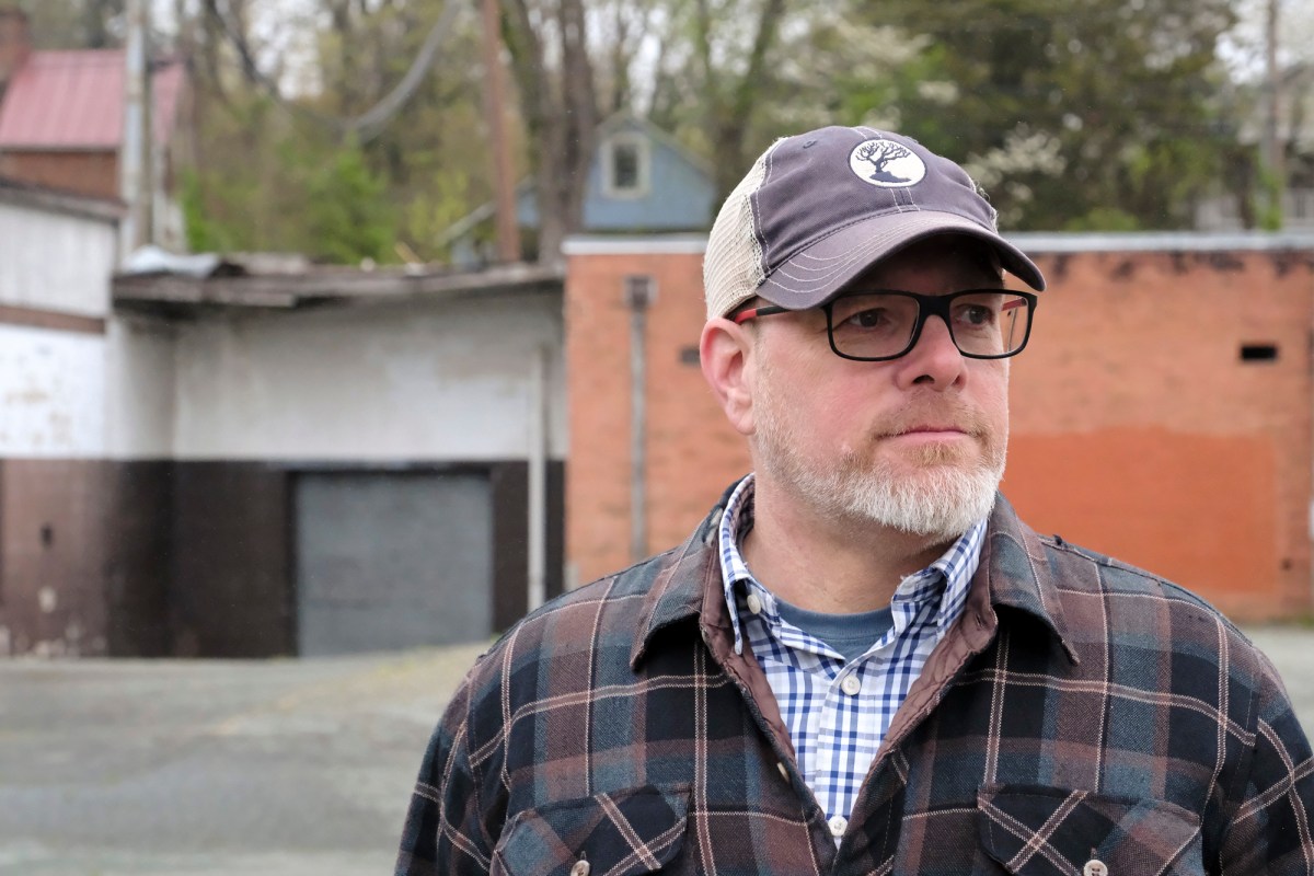 A close-up photo of a man wearing glasses, a hat, and a flannel jacket. In the background is an aging brick building with a sagging roof.