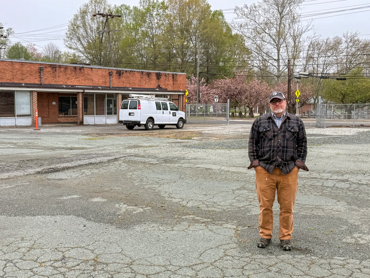A man wearing glasses, a baseball cap and a flannel jacket stands in a parking lot with cracked pavement. Behind him is an older brick building with a sagging roof and a van with ladders and other equipment parked beside it.