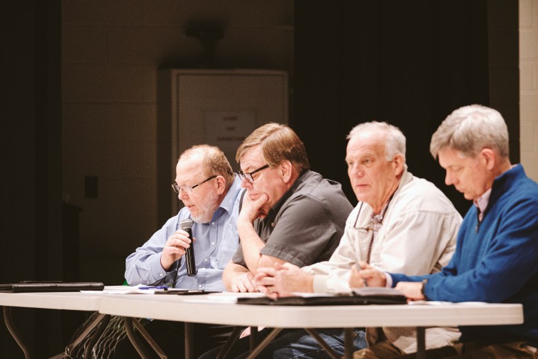 Four men seated at a long table taking notes and consulting with papers on the table. One man is holding a microphone and speaking.