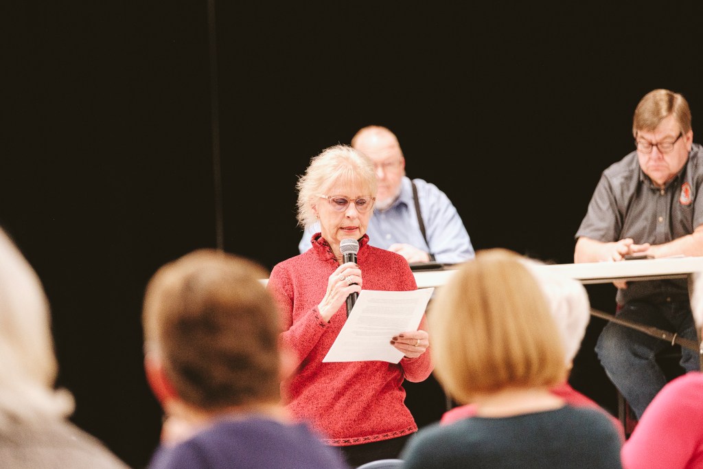 A woman with glasses holds a microphone and speaks while reading off of a piece of paper she is holding in her hand. She is facing a crowd of people who are seated, and there are two men visible seated at a long table behind her.